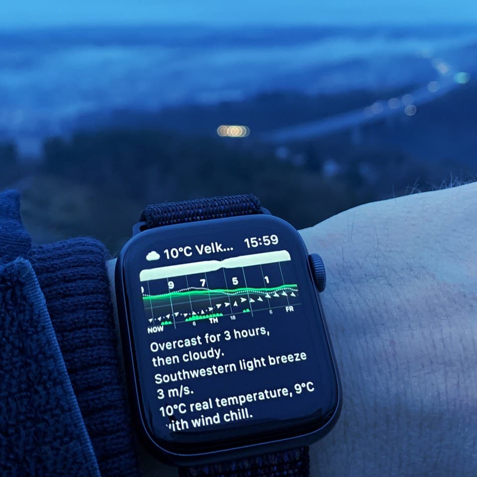 Hiker wearing Apple Watch displaying Weathergraph weather complication with temperature graph and sunshine indicators, photographed from mountain lookout tower